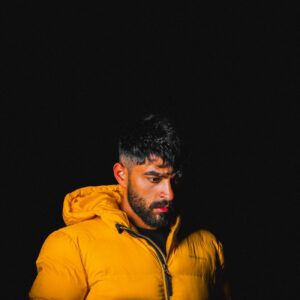 Dramatic portrait of a man with beard wearing a yellow jacket against a dark background.