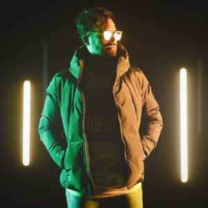 A man in fashionable eyewear and jacket stands amidst neon lights, exuding modern style.
